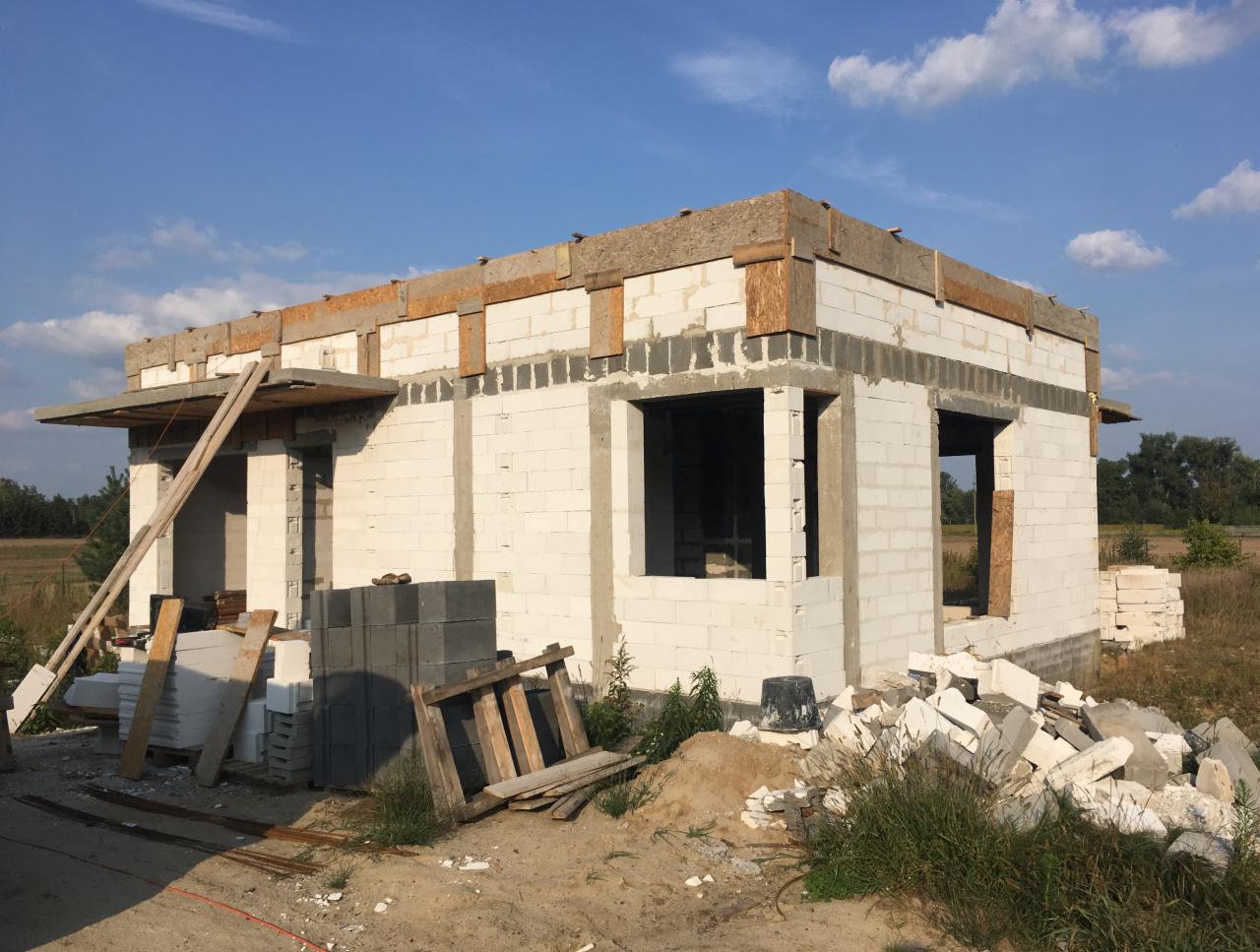 Surowy stan budynku jednorodzinnego z białych bloczków, widoczne otwory okienne i drzwiowe, drewniane elementy konstrukcyjne pod dachem, teren budowy z gruzem i materiałami budowlanymi, błękitne...