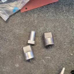 Trzy metalowe elementy z gwintem na szorstkiej, ciemnej powierzchni, obok woreczek z niebieskimi śladami i kawałek papieru ściernego.