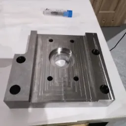 Szara, metalowa forma z wyfrezowanymi rowkami i okrągłym otworem pośrodku, leżąca na białym stole, z pudełkiem wierteł i czerwonym organizerem narzędzi w tle.