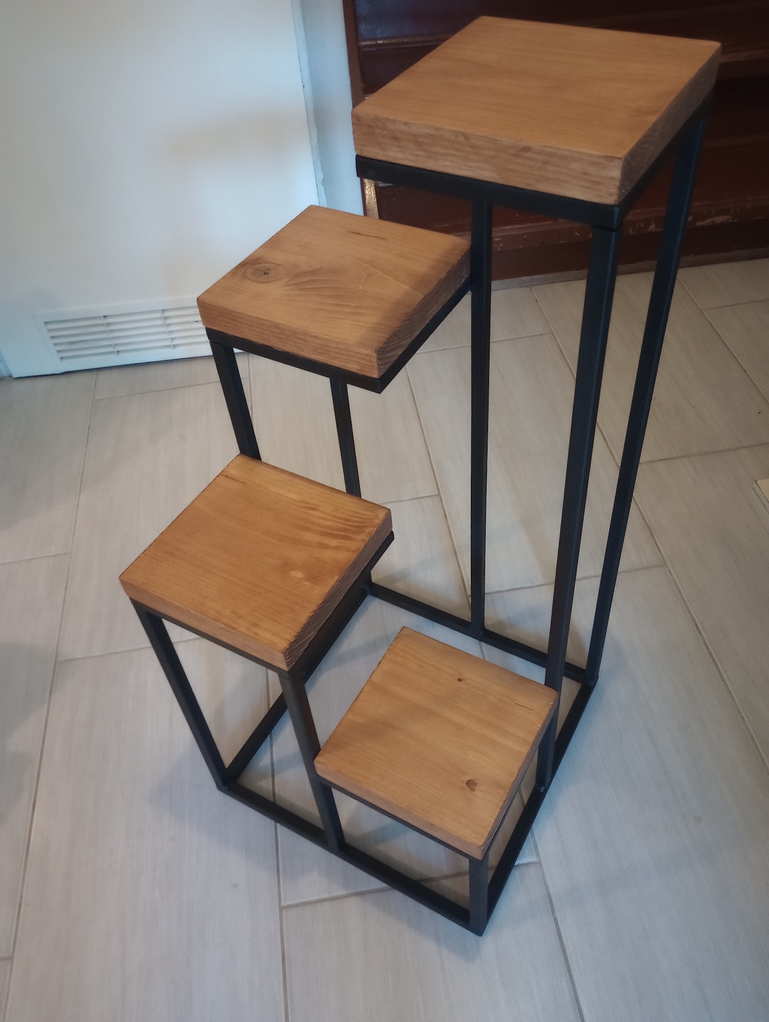 Metalowo-drewniany kwietnik schodkowy z trzema poziomami, na jasnej podłodze. Minimalistyczny design, czarne metalowe ramy i drewniane blaty.