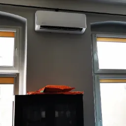 Biały klimatyzator Rotenso zamontowany na szarej ścianie w mieszkaniu w Bezrzeczu, widoczny nad czarnym barkiem z pomarańczową serwetą, pomiędzy dwoma oknami z roletami.