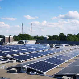 Panele fotowoltaiczne na dachu budynku przemysłowego, widoczne elementy montażowe i klimatyzatory, tło z zielenią i błękitnym niebem z chmurami.