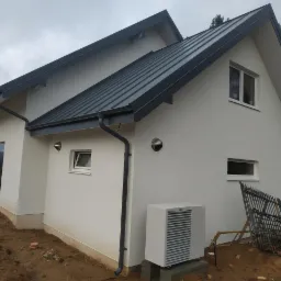 Nowoczesny dom z metalowym dachem i pompą ciepła Vaillant na zewnątrz, widok z boku w pochmurny dzień.