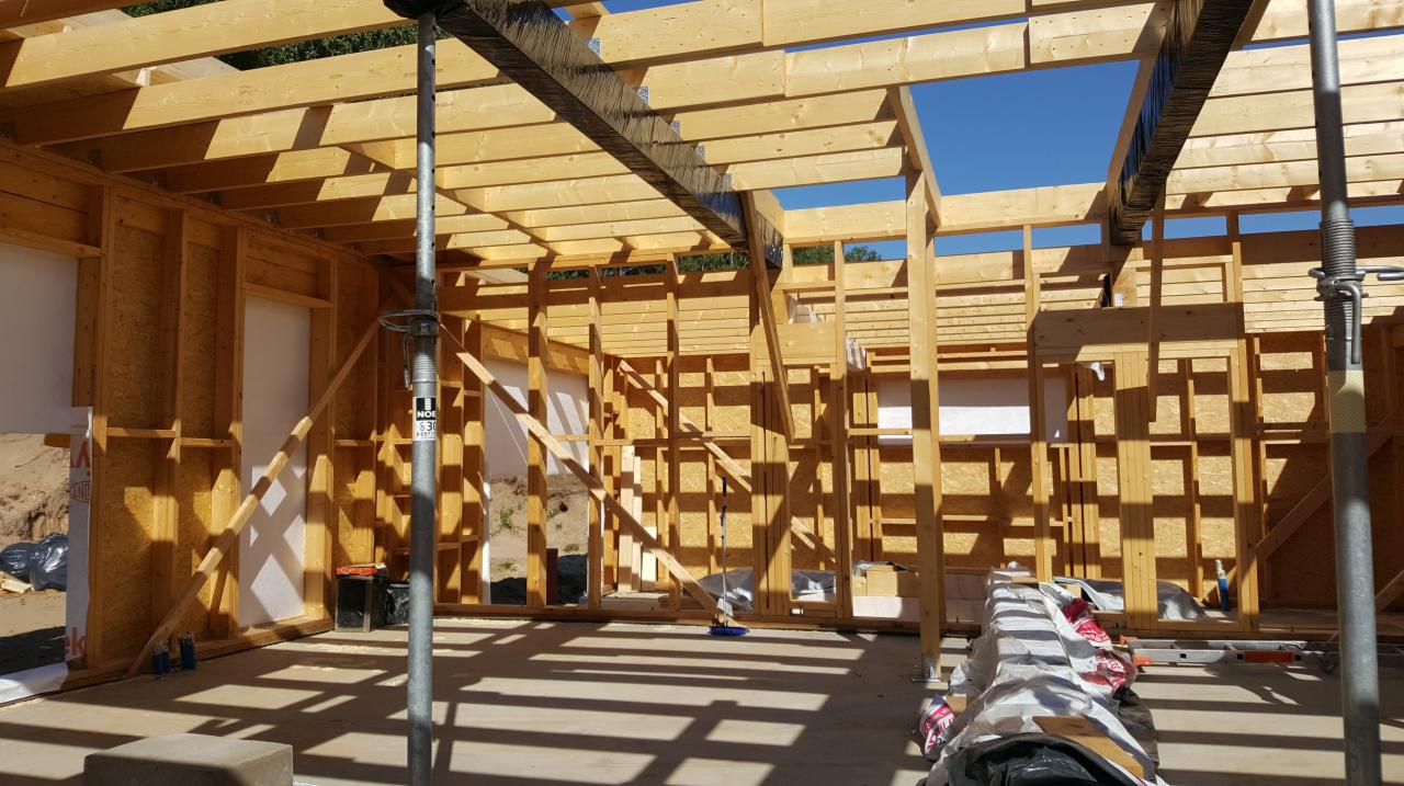 Wnętrze domu w trakcie budowy z drewnianą konstrukcją szkieletową, widoczne podpory stalowe i materiały budowlane na podłodze.