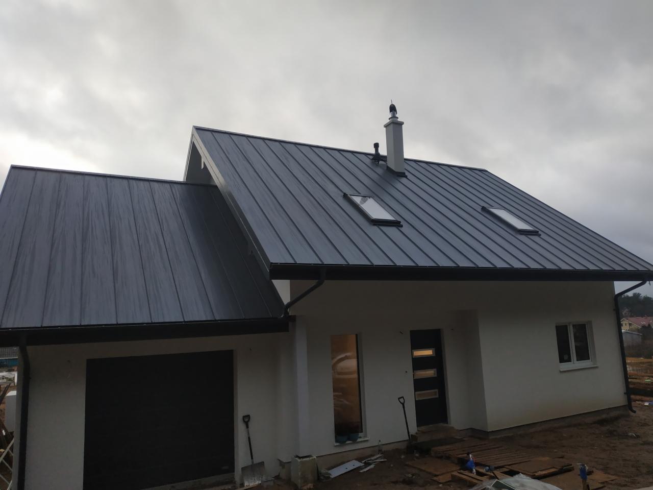 Nowoczesny dom jednorodzinny z ciemnoszarą blachodachówką na dachu, widoczne okna dachowe i komin, z garażem po lewej stronie, szpadel oparty o ścianę obok bramy garażowej.