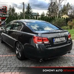 Ciemnoszary Lexus GS430 po detailingu, widok z tyłu na tle kostki brukowej i ogrodowej zieleni, z widocznym logo firmy detailingowej Domowy Auto Detailing Lublin.