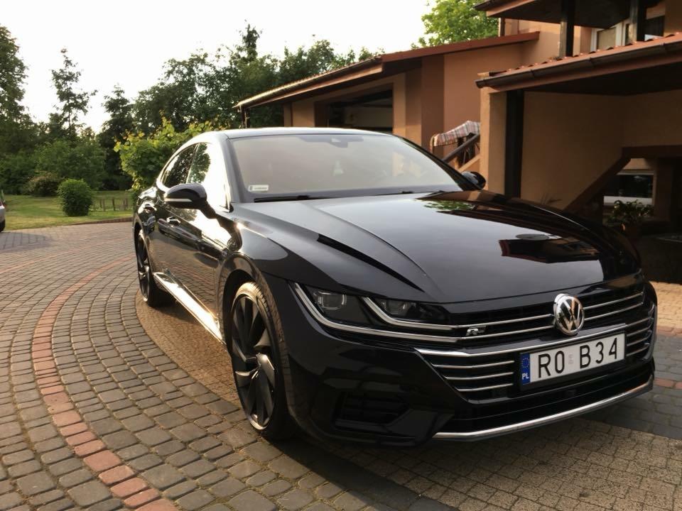 Błyszczący, czarny Volkswagen Arteon zaparkowany na brukowanym podjeździe przed domem w słoneczny dzień.