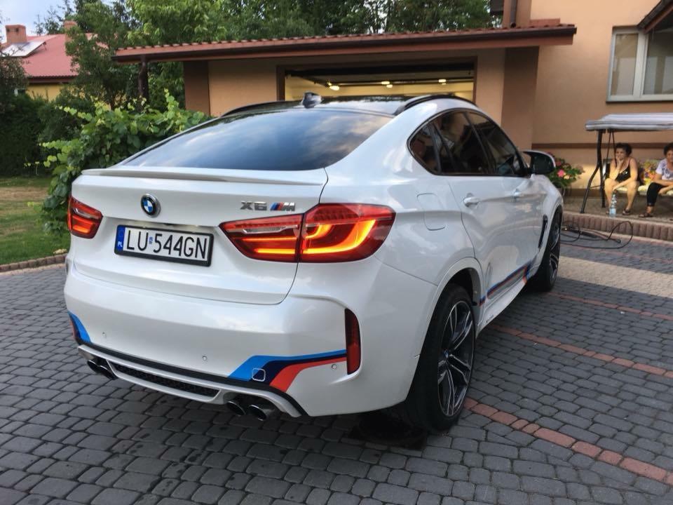 Białe BMW X6M zaparkowane na brukowanej nawierzchni przed budynkiem, widoczne detale jak tablica rejestracyjna, tylne światła, emblematy i charakterystyczne pasy w kolorach BMW Motorsport.