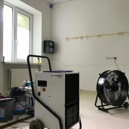 Pokój w trakcie osuszania, z widocznym osuszaczem Trotec i wentylatorem przemysłowym, ślady po taśmie malarskiej na ścianie, okno z grzejnikiem.