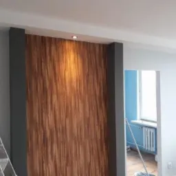 Ściana z drewnianą okładziną pionową, oświetlona punktowo, z szarą ramą po bokach, w tle widoczny fragment pokoju z drabiną i otwartym przejściem do innego pomieszczenia z niebieskimi ścianami...