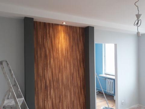 Ściana z drewnianą okładziną pionową, oświetlona punktowo, z szarą ramą po bokach, w tle widoczny fragment pokoju z drabiną i otwartym przejściem do innego pomieszczenia z niebieskimi ścianami...