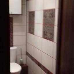 Kermaz - Wnętrze małej łazienki po remoncie: białe i brązowe płytki z cienkimi czerwonymi liniami, toaleta z białą szafką nad nią, różowy dywanik na podłodze.