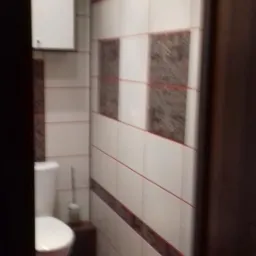 Wnętrze małej łazienki po remoncie: białe i brązowe płytki z cienkimi czerwonymi liniami, toaleta z białą szafką nad nią, r&oacute;żowy dywanik na podłodze.