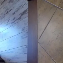 Połączenie podłogi z paneli imitujących bielone drewno oraz beżowych płytek ceramicznych, rozdzielonych brązową listwą przypodłogową, widok z góry w słabym oświetleniu.