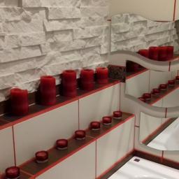 Kermaz - Ściana w łazience wykończona dekoracyjną białą cegłą, z półkami na których ustawione są czerwone świece różnej wielkości, odbijające się w falistym lustrze.