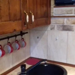 Fragment kuchni z drewnianymi szafkami, czarnym zlewem i rzędem czerwonych kubków zawieszonych na relingu, ściany wyłożone płytkami imitującymi cegłę i beżowymi płytkami.