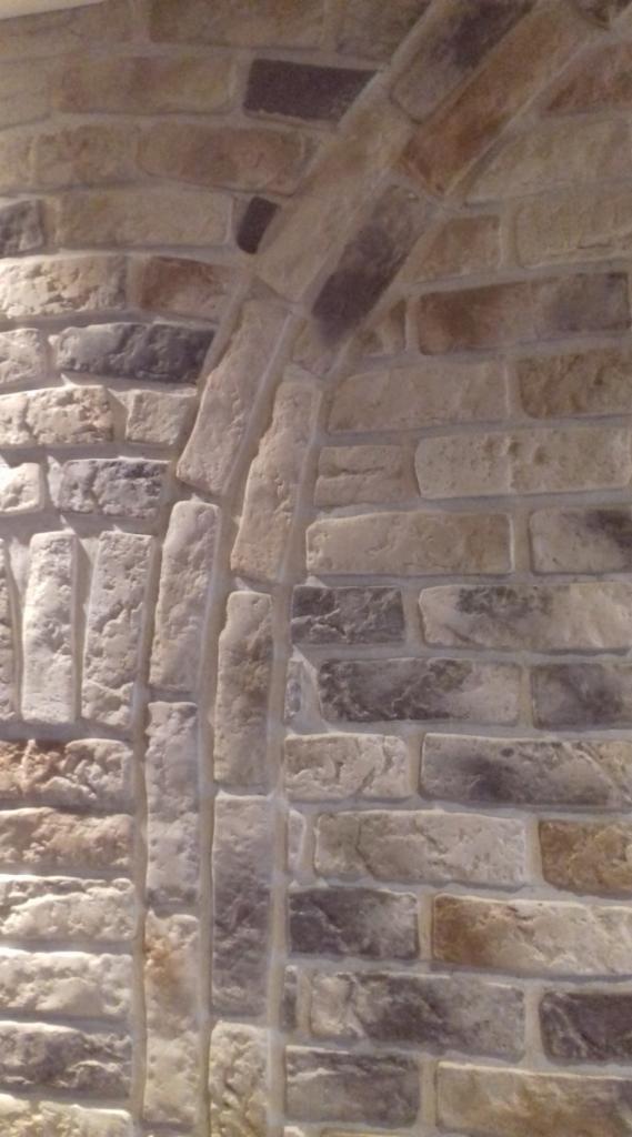 Ściana z dekoracyjnej cegły w odcieniach beżu i szarości, ułożona w łukowatym kształcie, imitująca starą architekturę.