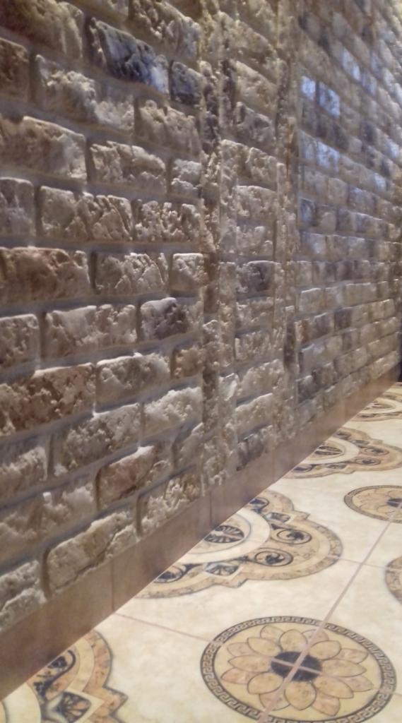 Ściana wykończona dekoracyjną cegłą w odcieniach beżu i brązu, z widoczną fakturą i nierównościami, połączona z podłogą wyłożoną płytkami w stylu vintage z motywem kwiatowym.