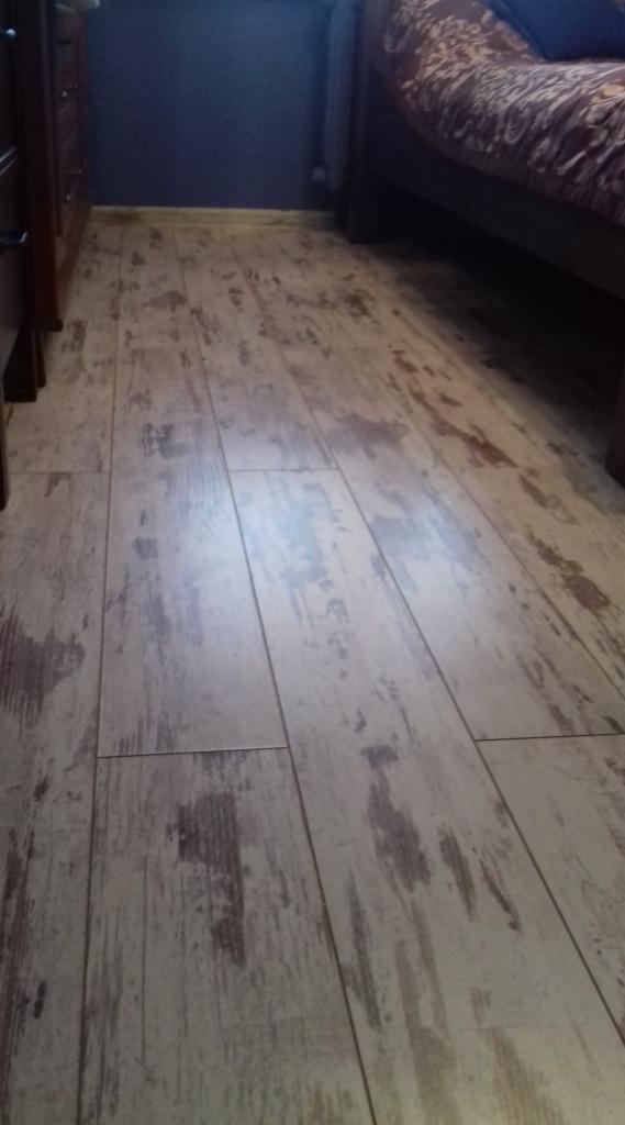 Ułożone panele podłogowe imitujące stare, bielone drewno, widoczne fragmenty mebli w tle.