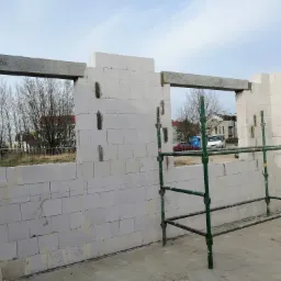 Wznoszenie ścian z białych bloczków, widoczne nadproża okienne, fragment rusztowania, w tle zabudowa jednorodzinna.