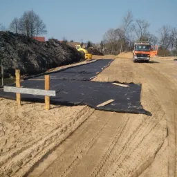 Przygotowanie terenu pod budowę: rozłożona czarna geowłóknina na piasku, widoczne ślady opon ciężarówki i koparka w tle, drewniane paliki stabilizujące grunt.