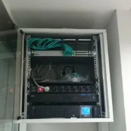 Otwarta szafa rack z zainstalowanym sprzętem sieciowym: patch panel, switch, listwa zasilająca, UPS z wyświetlaczem LCD. Widoczne okablowanie i częściowo przezroczyste drzwi szafy.