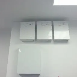 Trzy białe skrzynki alarmowe z schematami na ścianie w biurze, oraz czwarta mniejsza skrzynka pod nimi. Oświetlenie sufitowe LED.