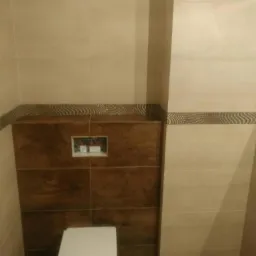 Nowoczesna toaleta podwieszana z brązową zabudową i beżowymi płytkami, wykończona dekoracyjnym pasem mozaiki.