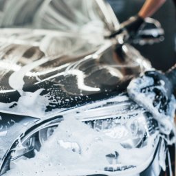 Mega Myjnia - Czyszczenie reflektora czarnego auta za pomocą gąbki. Widoczna piana z detergentu i ręka osoby w czarnej rękawicy. Detailing samochodu w zbliżeniu.