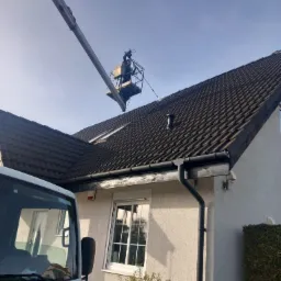 Pracownik na podnośniku koszowym czyści dach z ciemnej dachówki. Widoczny fragment elewacji domu i firmowy samochód z adresem strony internetowej.
