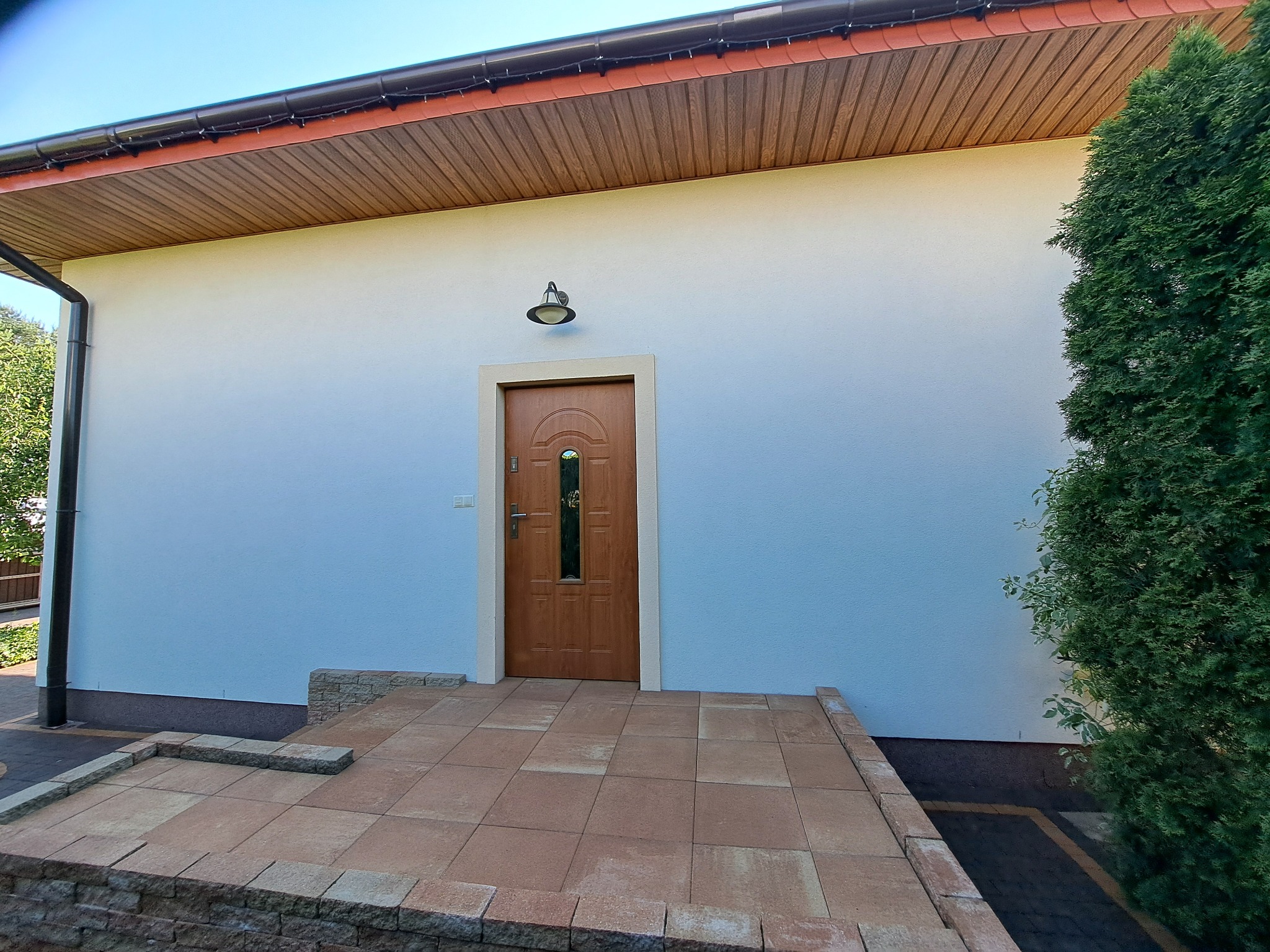 Wejście do domu z brązowymi drzwiami, jasną elewacją i kostką brukową w odcieniach brązu, oświetlone lampą zewnętrzną.