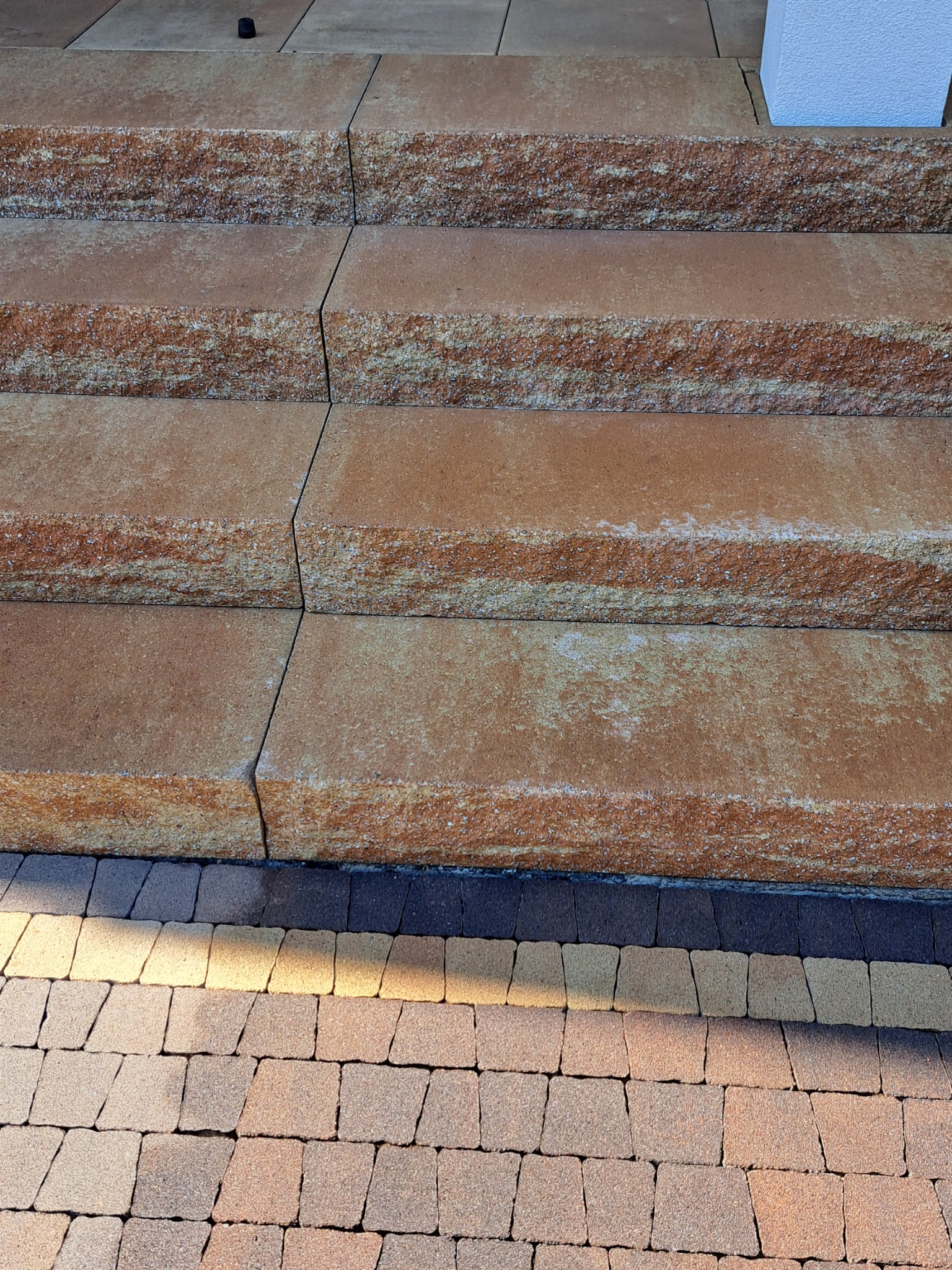 Schody zewnętrzne z kostki brukowej w odcieniach brązu i beżu, prowadzące do budynku z białym elementem elewacji, widoczne z bliska z perspektywy lekko od dołu.