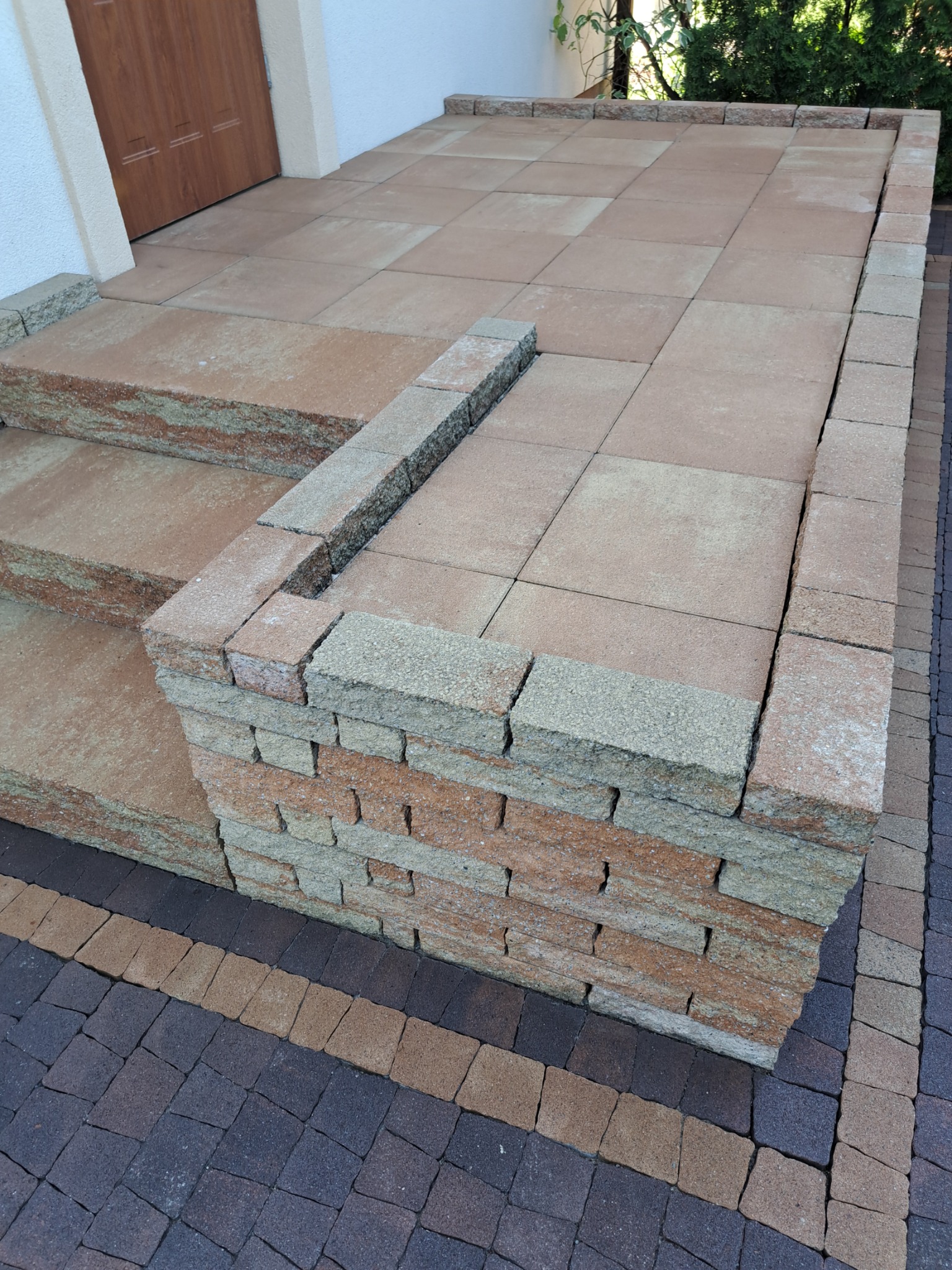 Ułożone schody wejściowe z kostki brukowej w odcieniach beżu i brązu, z widocznym fragmentem chodnika z ciemnej kostki brukowej w tle.