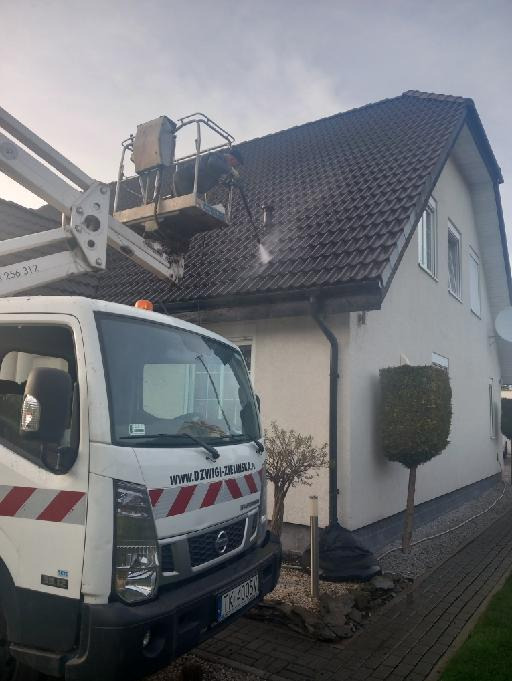 Mycie dachu z podnośnika koszowego. Pracownik czyści dach z zanieczyszczeń za pomocą myjki ciśnieniowej. Biała elewacja budynku.