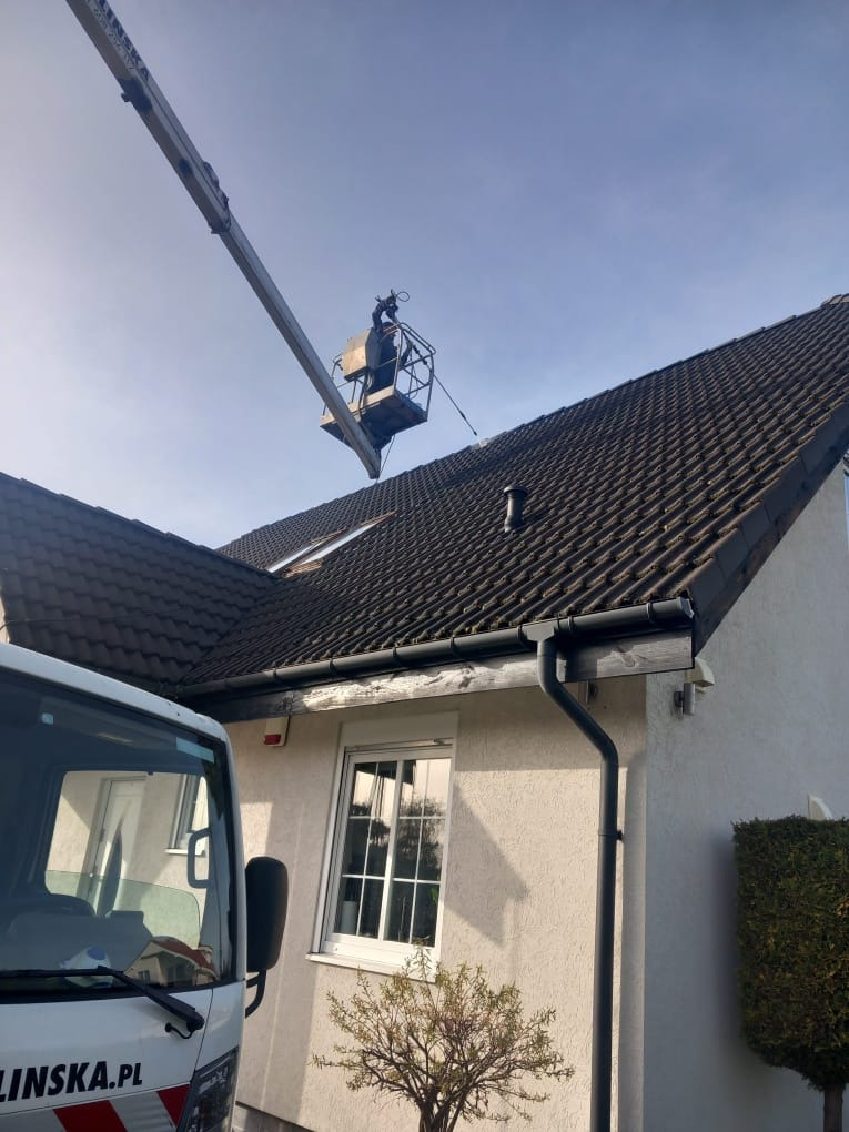 Pracownik na podnośniku koszowym czyści dach z ciemnej dachówki. Widoczny fragment elewacji domu i firmowy samochód z adresem strony internetowej.