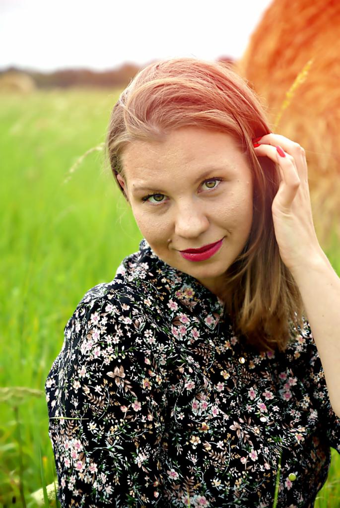 Portret kobiety w kwiecistej bluzce na tle zielonej łąki i stogu siana, z ręką we włosach i delikatnym uśmiechem.