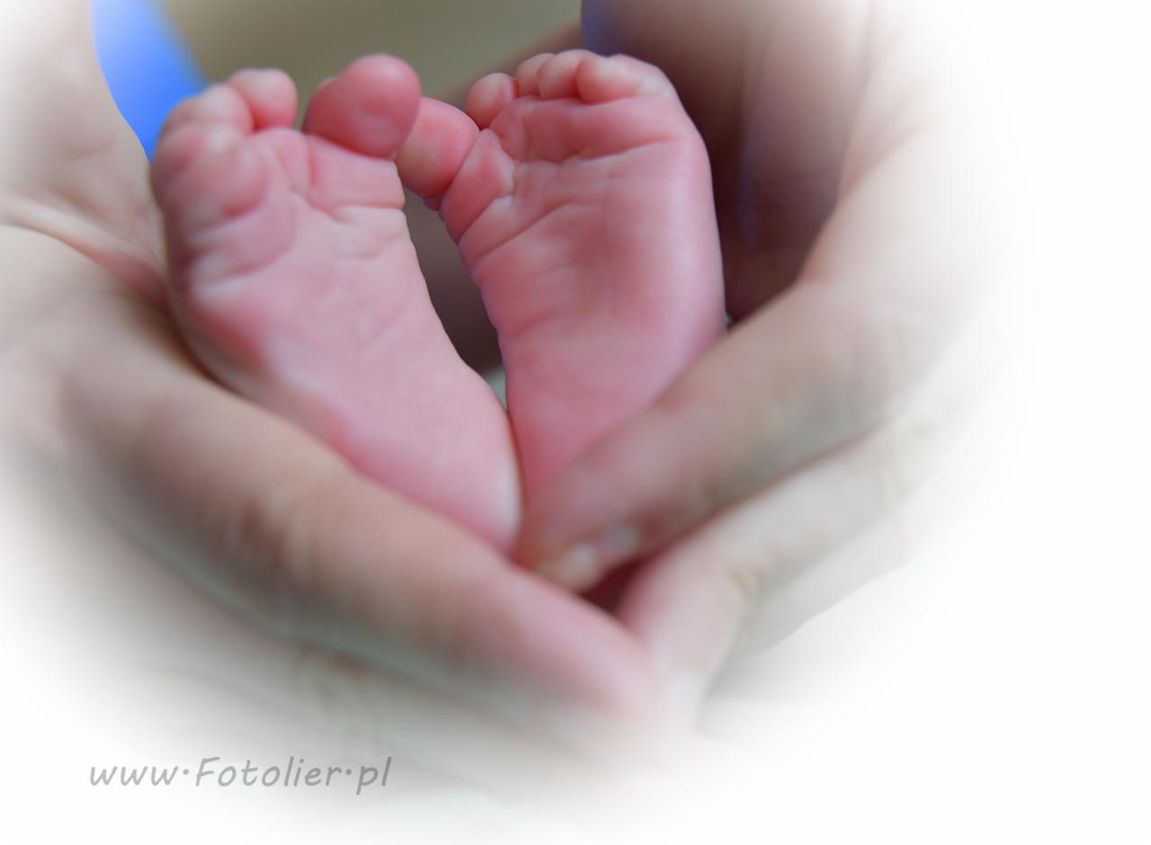 Drobne stópki noworodka, delikatnie trzymane w dłoniach dorosłego, z widocznym logo fotografa w dolnej części kadru.