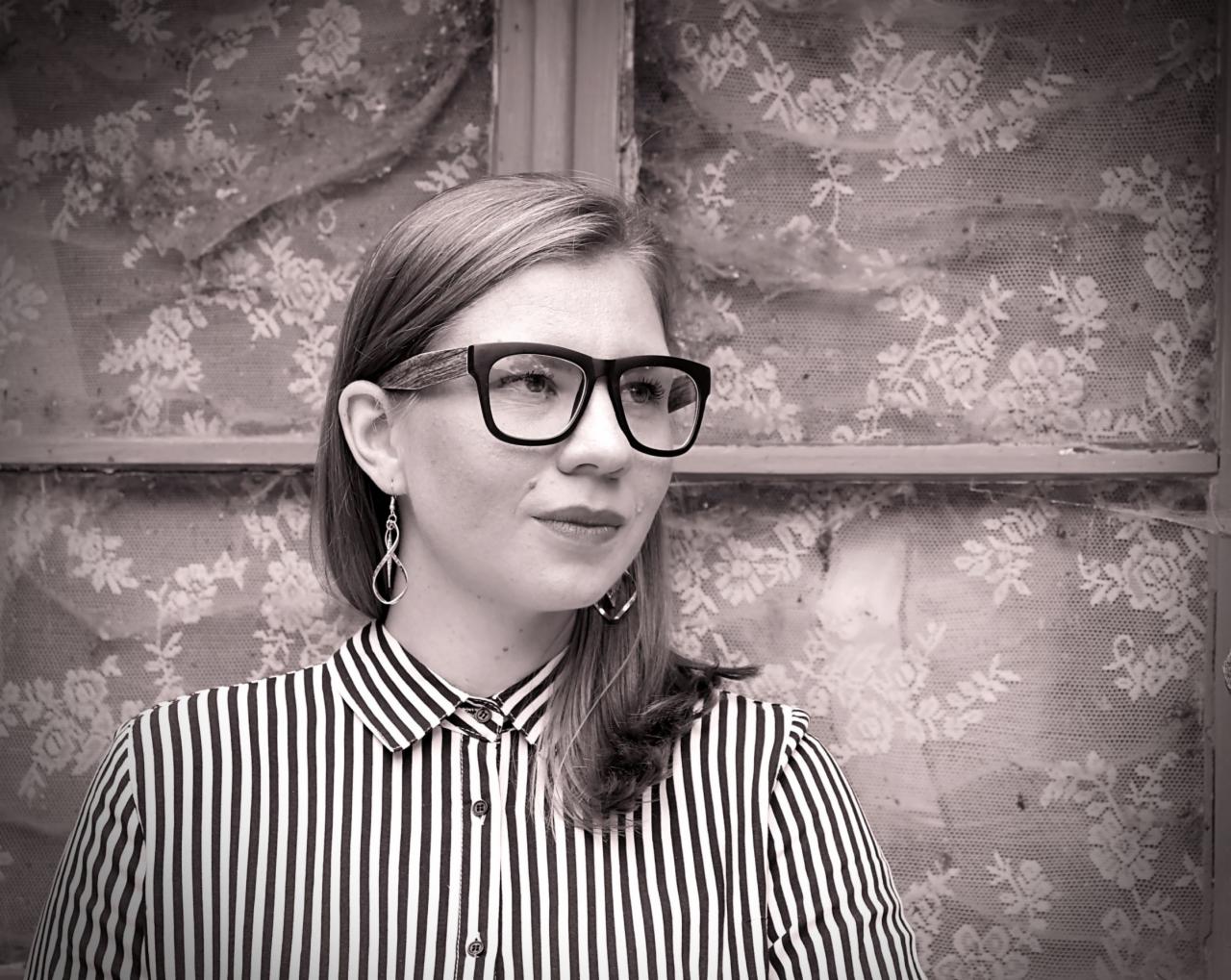 Portret kobiety w okularach z czarnymi oprawkami, w bluzce w czarno-białe paski, stojącej na tle okna z koronkową zasłoną, w sepii.