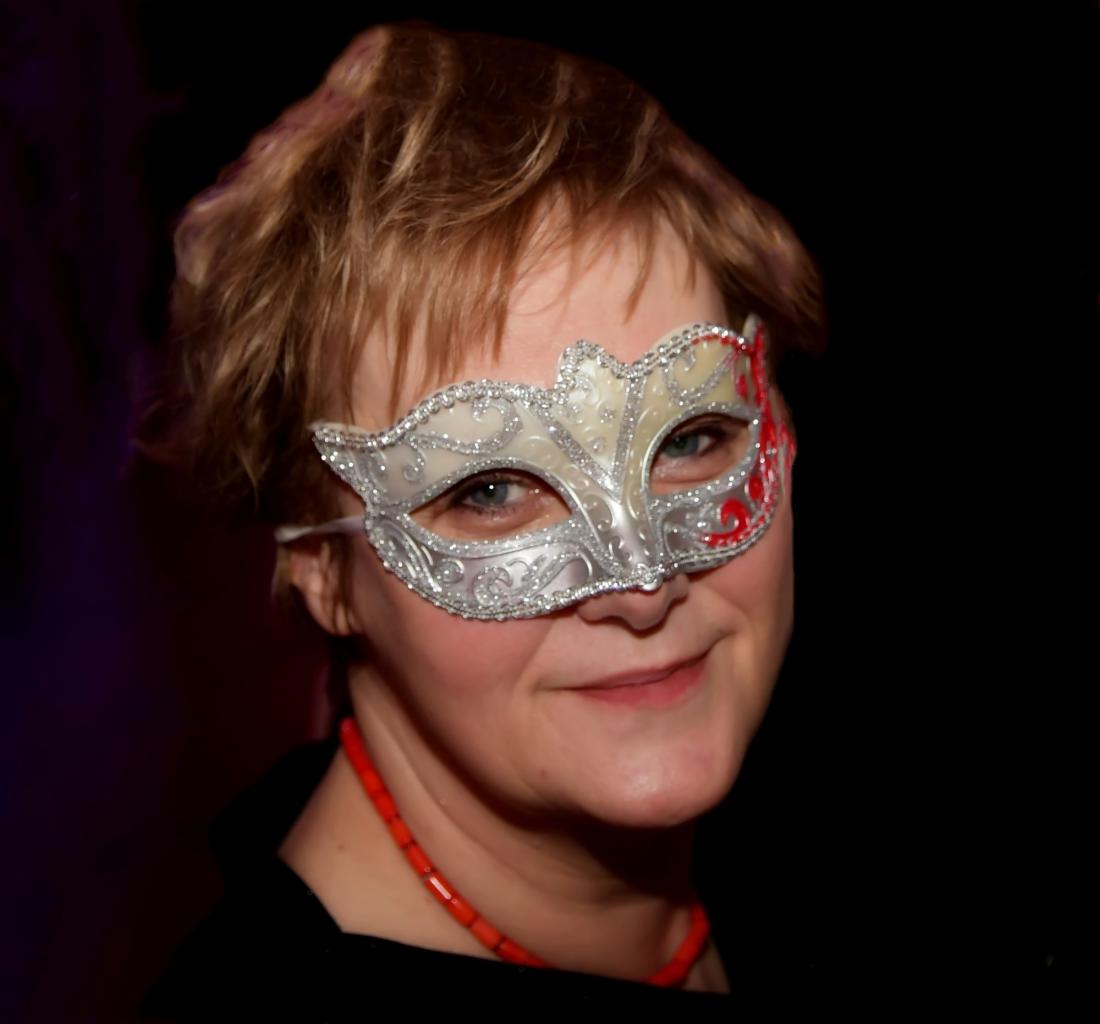 Portret kobiety w srebrnej masce weneckiej z czerwonym akcentem, noszącej koralowy naszyjnik i czarną bluzkę, na ciemnym tle.