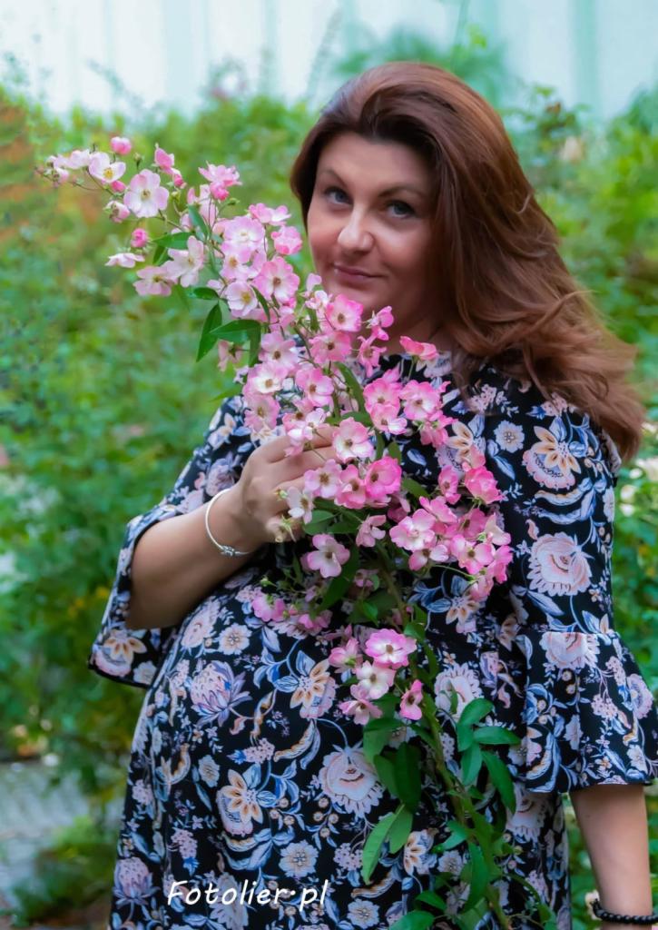 Kobieta w kwiecistej sukience pozuje z gałązką kwitnącej róży w ogrodzie, uśmiechając się delikatnie w stronę obiektywu.