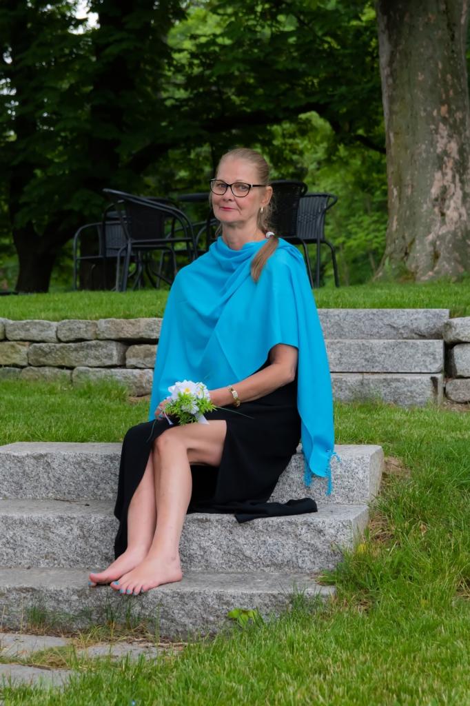 Kobieta w okularach, czarnej sukience i turkusowym szalu siedzi boso na kamiennych schodach, trzymając mały bukiet kwiatów na tle zieleni.