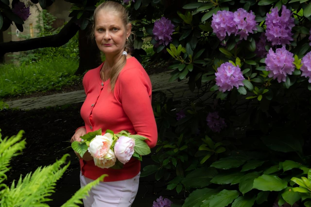 Kobieta w koralowej bluzce i białych spodniach trzyma bukiet różowych i białych piwonii na tle zieleni i fioletowych rododendronów.