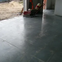 Wypoziomowana, świeżo wylana, ciemnoszara posadzka z widocznym śladem po zacieraczce mechaniczej, w tle sterta piasku i fragment ogrodzenia.