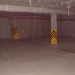 Rut-Bud - Surowa, betonowa przestrzeń z pomalowanymi na żółto słupami z czarnymi strzałkami, prawdopodobnie garaż podziemny lub hala przemysłowa, z widocznymi instalacjami pod sufitem.