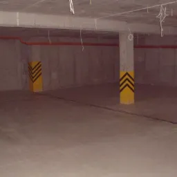 Surowa, betonowa przestrzeń z pomalowanymi na żółto słupami z czarnymi strzałkami, prawdopodobnie garaż podziemny lub hala przemysłowa, z widocznymi instalacjami pod sufitem.