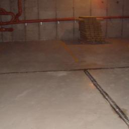 Rut-Bud - Szara, gładka posadzka w garażu podziemnym z widocznymi dylatacjami, pomalowanymi słupami ostrzegawczymi i instalacją pod sufitem.