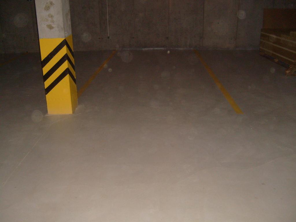 Gładka, szara posadzka w hali garażowej z żółtymi liniami wyznaczającymi miejsca parkingowe i słupem oznaczonym czarno-żółtymi pasami ostrzegawczymi.