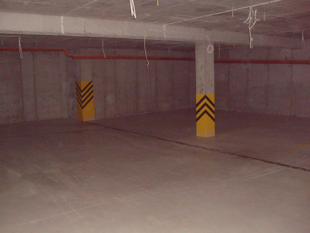 Surowa, betonowa przestrzeń z pomalowanymi na żółto słupami z czarnymi strzałkami, prawdopodobnie garaż podziemny lub hala przemysłowa, z widocznymi instalacjami pod sufitem.