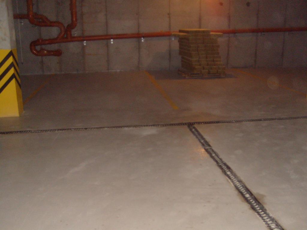 Szara, gładka posadzka w garażu podziemnym z widocznymi dylatacjami, pomalowanymi słupami ostrzegawczymi i instalacją pod sufitem.