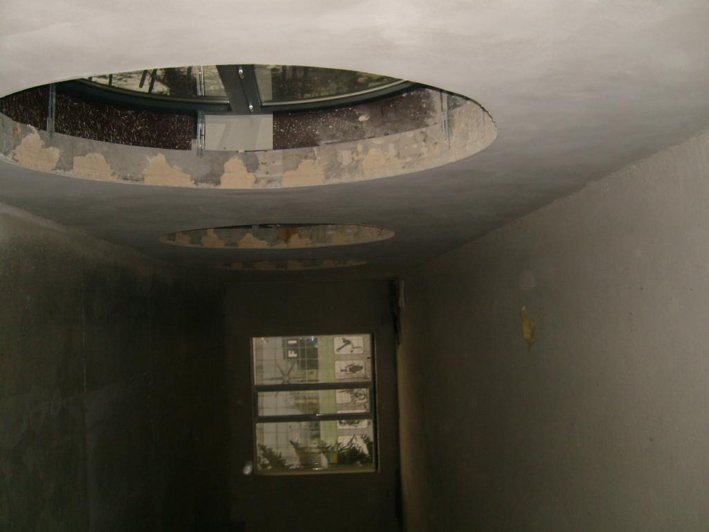 Wnętrze w trakcie prac wykończeniowych, widoczne otwory w suficie przygotowane pod instalacje, ściany pokryte szpachlą, okno z widokiem na budynek.